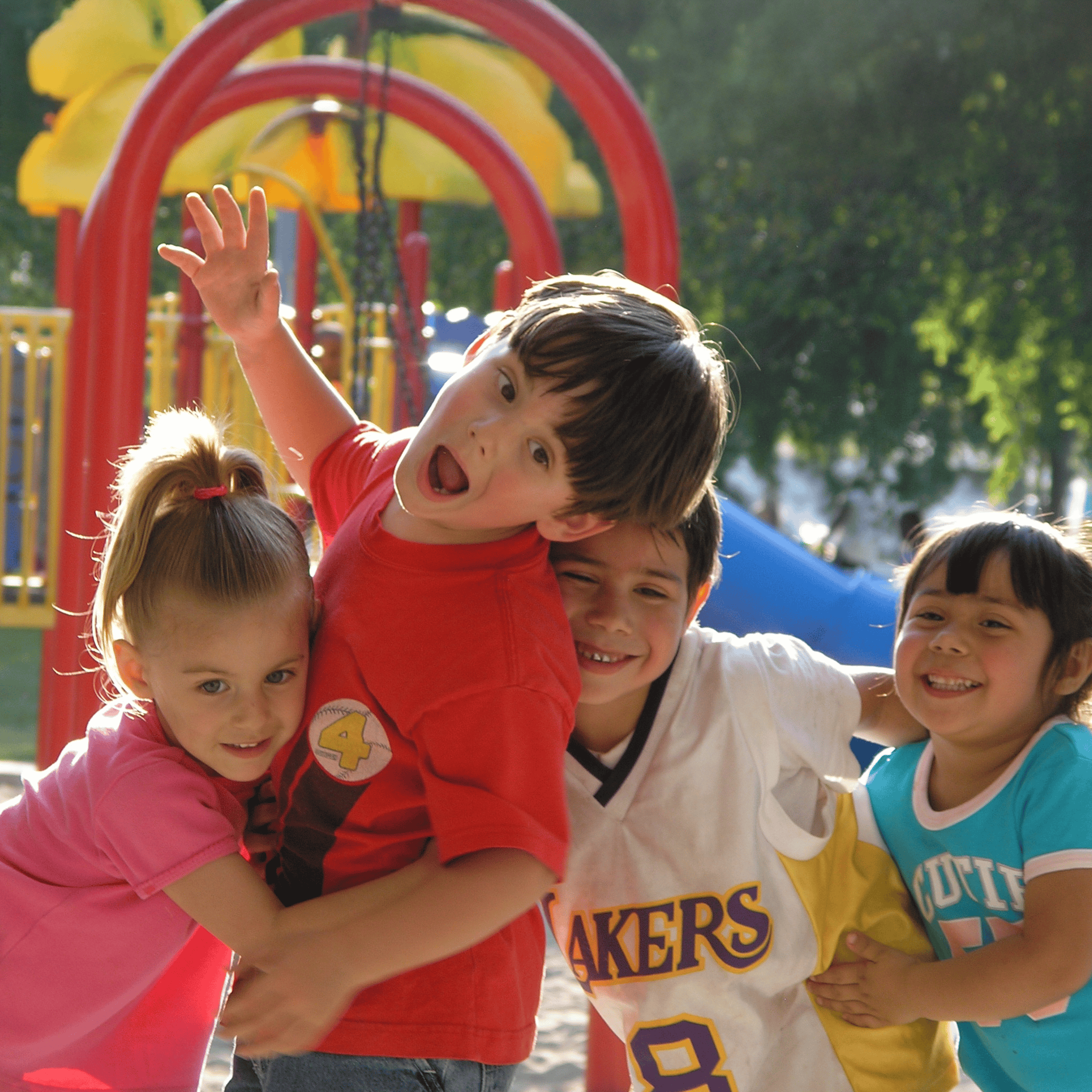 Four smiling children playing together at a colorful playground, one boy waving excitedly with arms raised