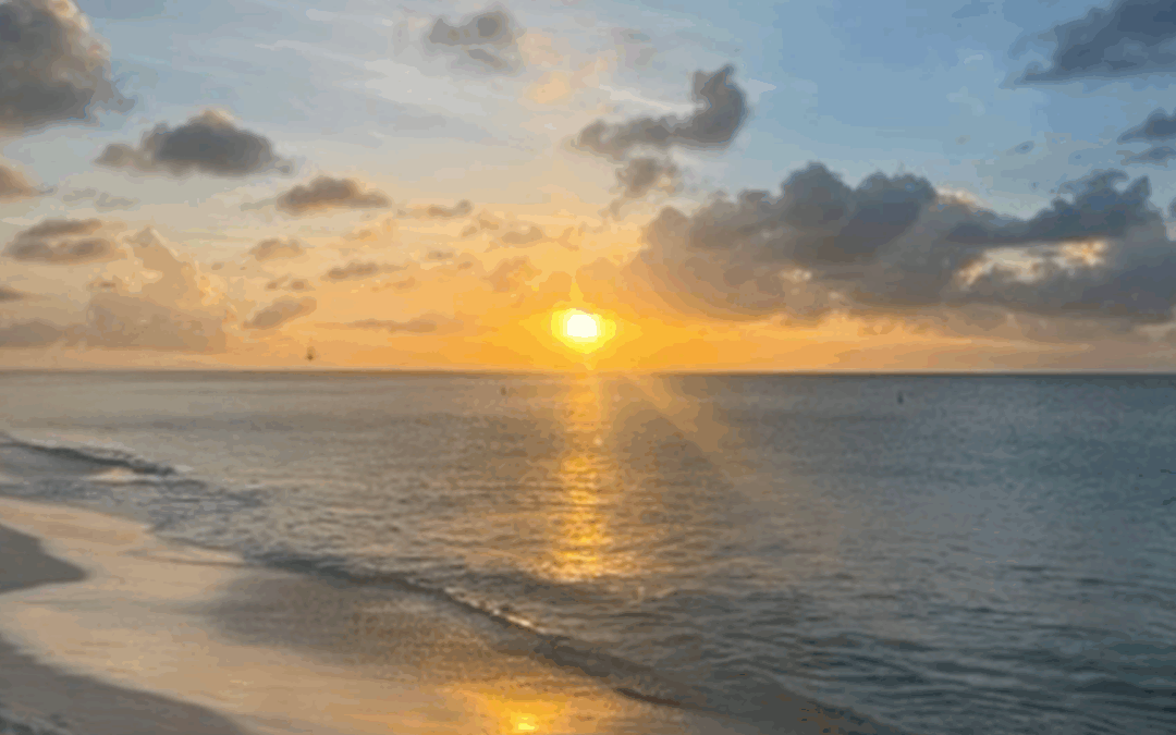 Golden sunset over calm ocean waters with sun reflecting on gentle waves and sandy shoreline under scattered clouds